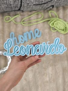 Light blue Tricotin shaping frame with the word Leonardo held in hand with knitted creations in the background